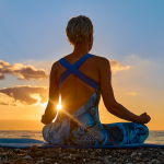Frau, die im Schneidersitz meditiert, an einem Strand bei Sonnenuntergang